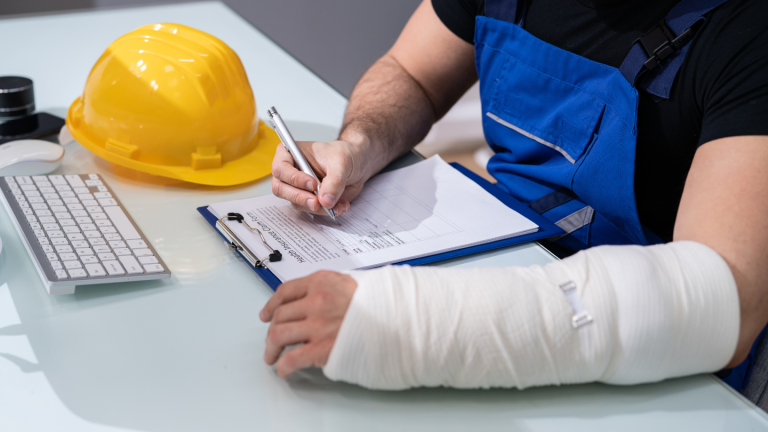 Man sits at a desk filling out paperwork with a construction hat on the next next to him and the left wrist is in a cast and bandaged up